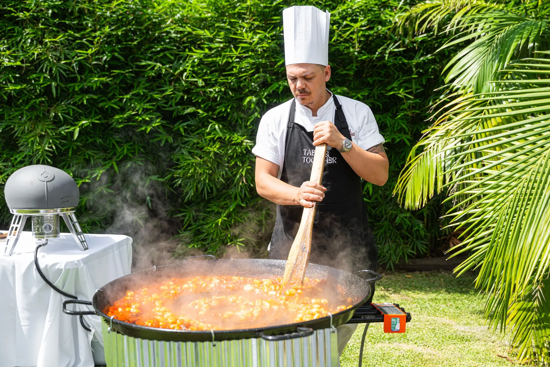 A chef in a tall white toque stirring a huge pan of tomato sauce and gnocchi at an outdoor cooking station, palm trees behind him