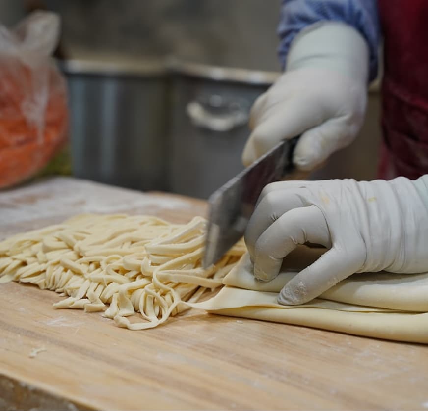 Gloved hands cutting fresh pasta dough into strands on a wooden board